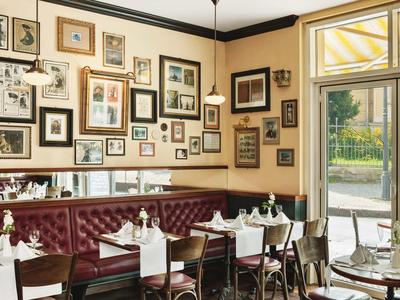 Gemütliches Café mit rot gepolsterten Sitzbänken, weißen Tischen, Stühlen und vielen gerahmten Bildern an der Wand.