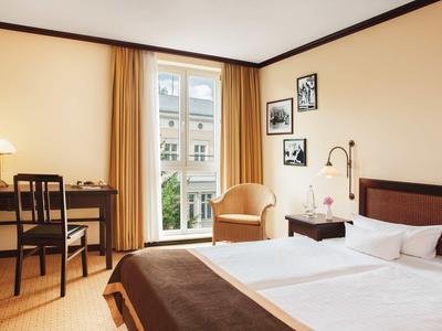 Helles Hotelzimmer mit Doppelbett, Stuhl, Schreibtisch und Blick auf ein Fenster mit Vorhängen.