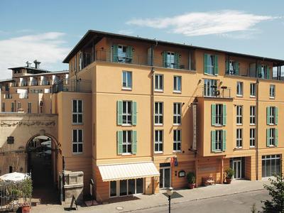 Gelbes Hotelgebäude mit blauen Fensterläden und Eingang unter dem Vordach in einer Stadtumgebung.