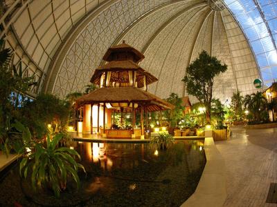 Ein exotisches Innenressort mit einem Pavillon, umgeben von Pflanzen und Wasser unter einer großen Glaskuppel.