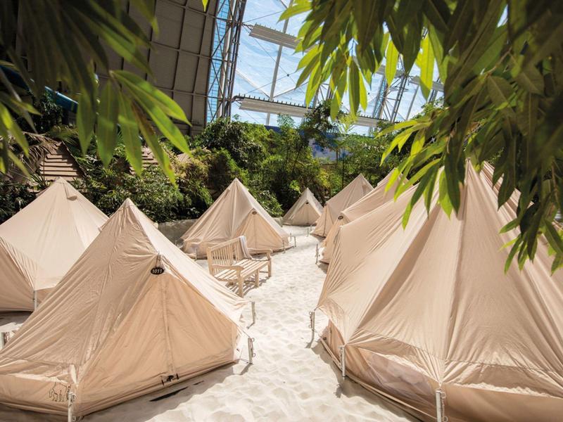 Mehrere sandfarbene Zelte stehen auf einem sandigen Boden unter einem Glasdach und grünen Pflanzen.