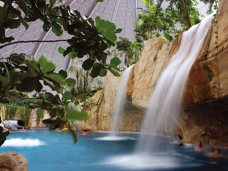 Innenpool mit Wasserfällen und Felsen unter begrüntem Dach in einem Hotel.