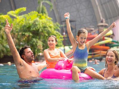 Family with two children playing in pool on pink flamingo float, surrounded by plants.