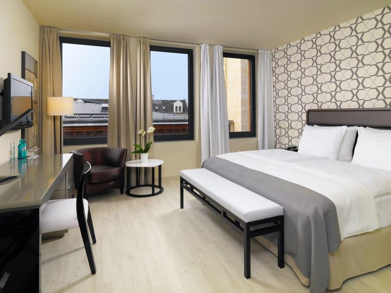 Habitación de hotel moderna con cama doble, escritorio, sillón y ventana con vista a edificios.