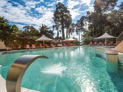 Grande piscina all'aperto con giochi d'acqua, lettini e ombrelloni in un hotel sotto un cielo azzurro.