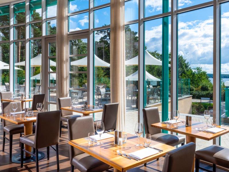 Ristorante luminoso con grandi finestre che si affacciano su terrazza e giardino con ombrelloni.