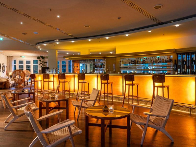 Interno di un bar d'hotel moderno con bancone illuminato e sedute in legno.