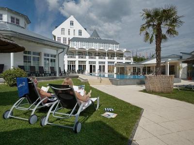 Zwei Personen entspannen auf Liegestühlen neben einem Pool in einem Hotel mit weißem Gebäude und Palmen.