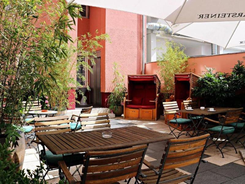 Gemütliche Terrasse mit Holztischen, Stühlen und grünem Pflanzenbewuchs in hellem Sonnenlicht.