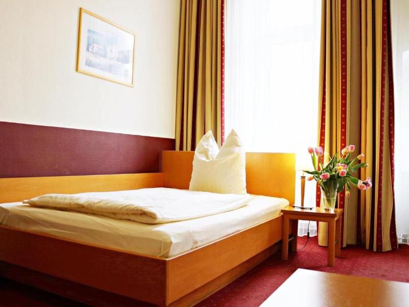 Schlichtes Einzelzimmer mit Holzbett, weißer Bettwäsche, roten Teppich und bunten Vorhängen.