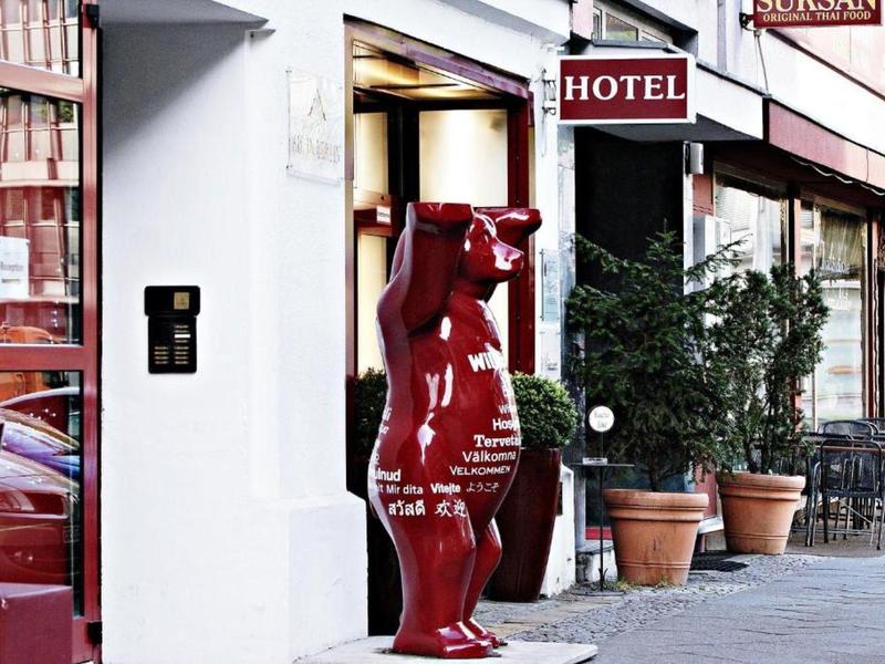 Rote Bärenfigur vor einem Hotel, neben blühenden Pflanzen in Töpfen auf dem Bürgersteig.