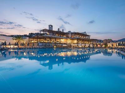 Een stijlvol hotel bij schemering met verlicht zwembad en rustig water, klassieke architectuur.