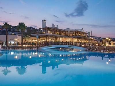 Abendlicher Blick auf einen Hotelpool mit beleuchteter Terrasse und reflektierendem Wasser.