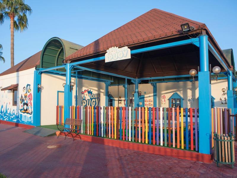 Kleurrijk gebouw met roodbruine dak en meerkleurige omheining onder blauwe lucht met palmbomen