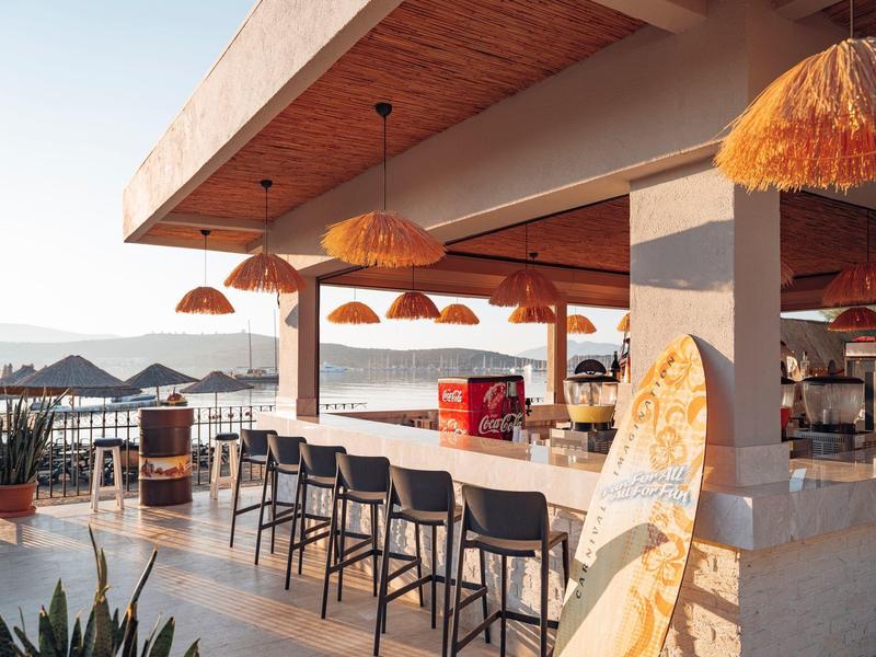 Open bar met krukken en strohoedlampen met uitzicht op zee en bergen op de achtergrond.