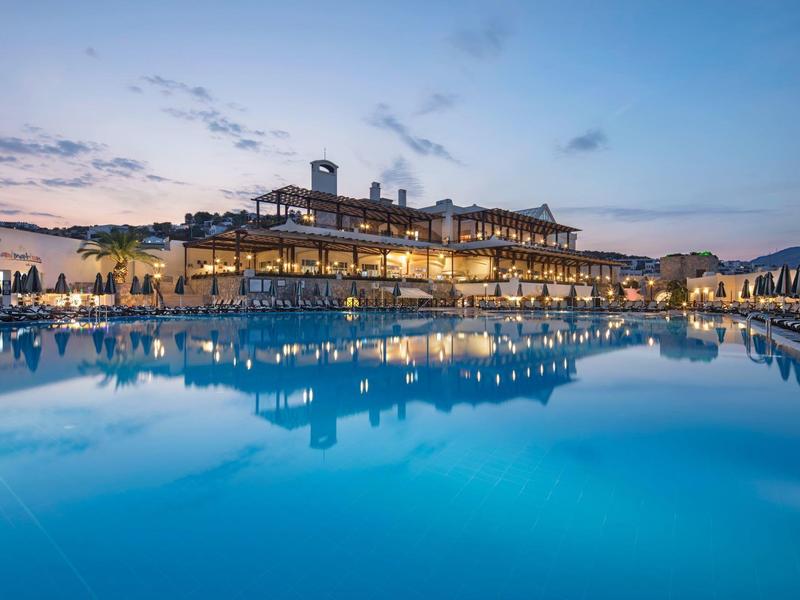 Een stijlvol hotel bij schemering met verlicht zwembad en rustig water, klassieke architectuur.