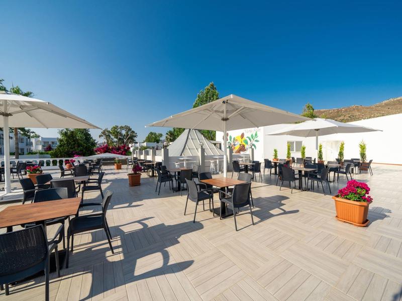 Słoneczny taras z stołami, krzesłami i parasolami, otoczony białymi ścianami.