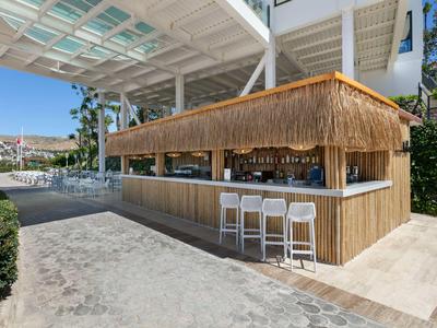 Moderner Strandbar mit Bambusverkleidung und weißen Barhockern unter einem weißen Dach.