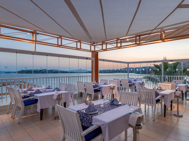 Zadaszony taras z nakrytymi stołami i widokiem na morze o zachodzie słońca.