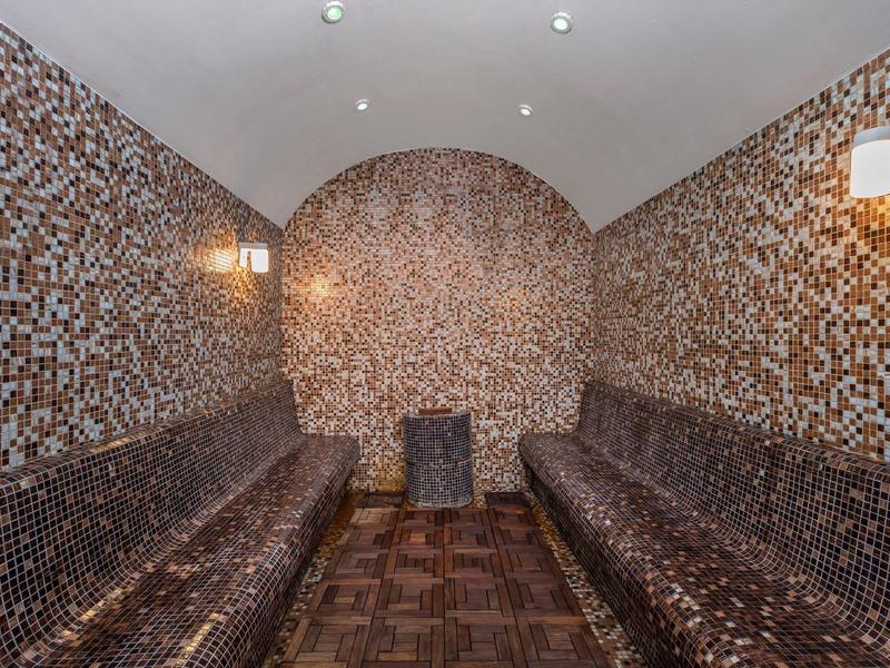 Wnętrze sauny z brązowymi mozaikowymi płytkami i dwoma długimi ławkami naprzeciwko siebie.