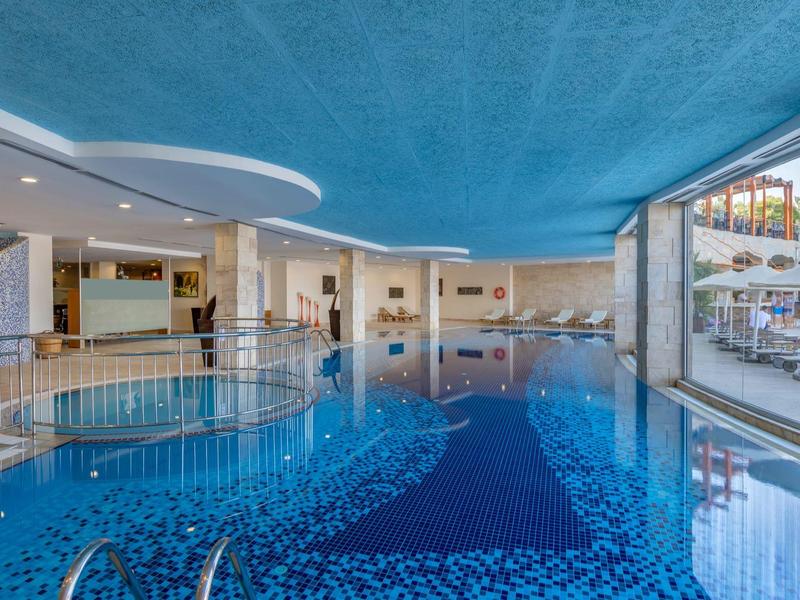 Basen kryty z niebieską wodą, jacuzzi, dużymi oknami i leżakami w hotelu.