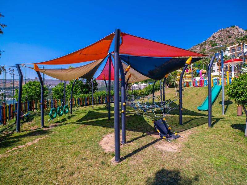 Kolorowy plac zabaw z huśtawkami, zjeżdżalnią i żaglami przeciwsłonecznymi na trawie.