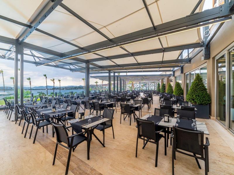 Duży zadaszony taras z nowoczesnymi czarnymi stołami i krzesłami, widok na teren zewnętrzny.