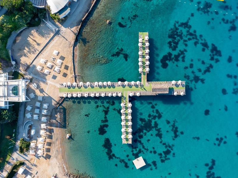 Widok z lotu ptaka na drewniany pomost z parasolami nad przejrzystą, niebieską wodą obok plaży z leżakami.