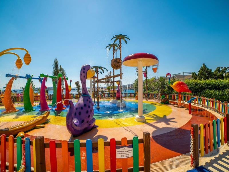 Kolorowy plac zabaw wodny z różnymi atrakcjami wodnymi, otoczony kolorowym ogrodzeniem pod niebieskim niebem.