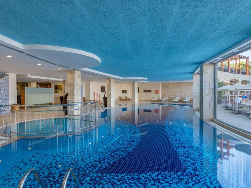 Duży kryty basen z niebieską wykładziną i szklaną ścianą z widokiem na leżaki.