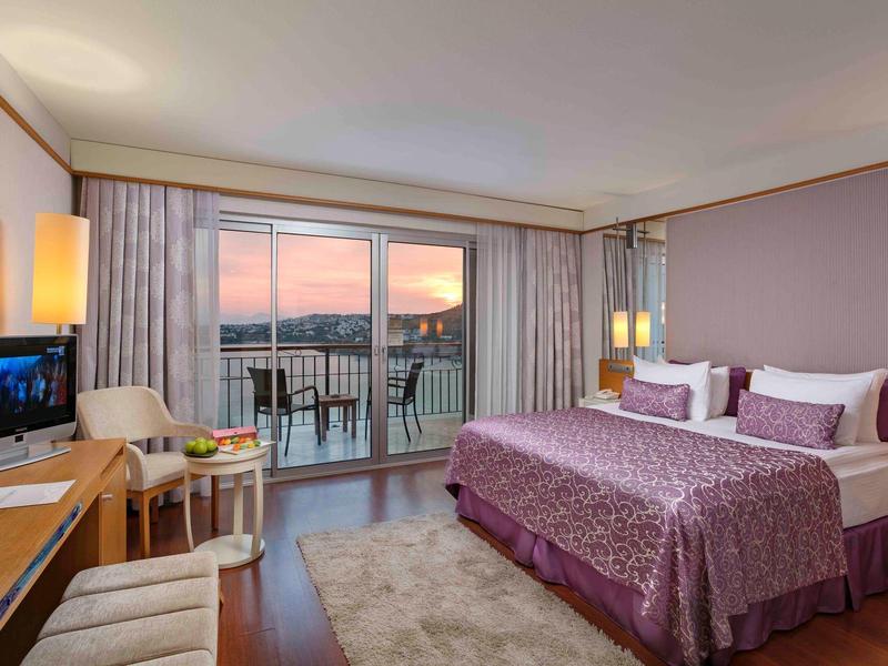 Sypialnia z balkonem, widokiem na morze o zachodzie słońca i fioletową narzutą w pokoju hotelowym.