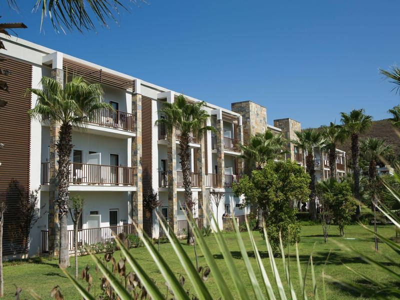 Modern hotelgebouw met drie verdiepingen en balkons, omringd door palmbomen en groen gras.