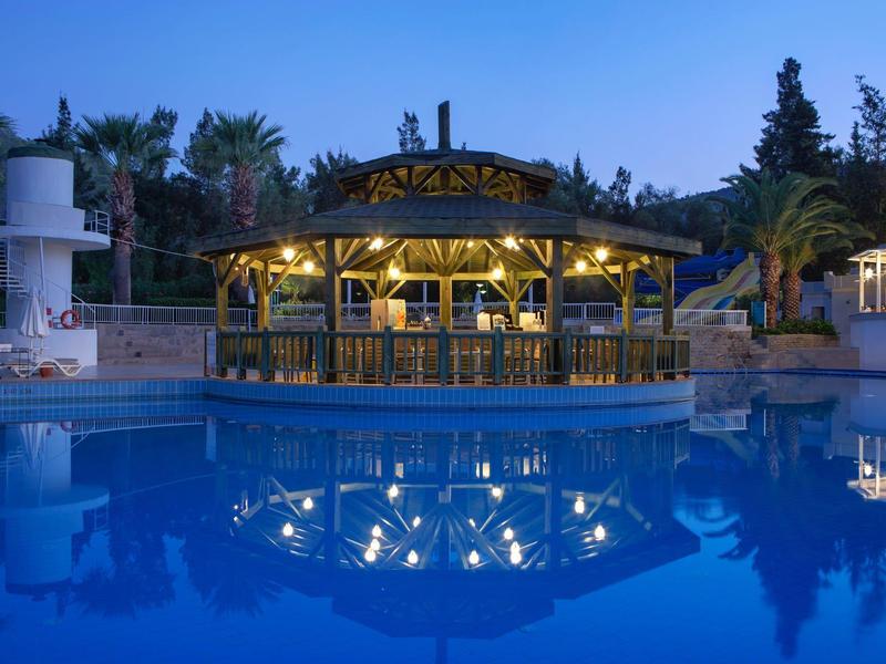 Poolbar verlicht bij schemering, reflecterend in rustig blauw water omringd door palmbomen.