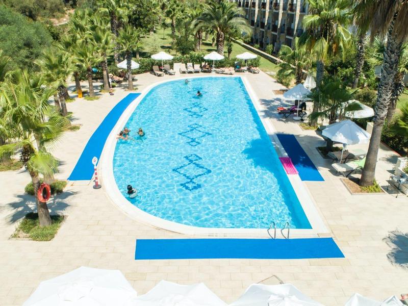 Buitenzwembad van hotel met ligstoelen, palmbomen en blauwe mozaïektegels in een zonnige omgeving.