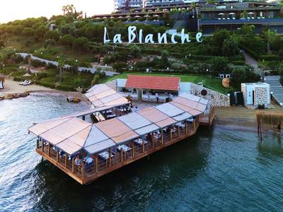Restaurant auf Holzplattform am Wasser mit grünen Hügeln und Hotel in Hintergrund.