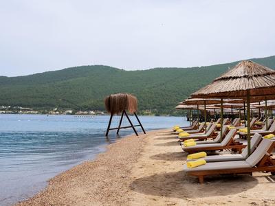 Strand mit Sonnenliegen, Sonnenschirmen und Meer mit grünem Hügel im Hintergrund.