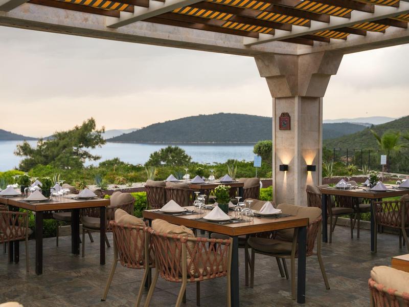 Área de restaurante abierta con mesas y sillas de madera con vistas al lago y montañas al atardecer.