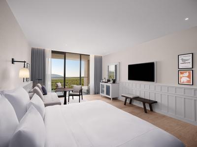 Helles Hotelzimmer mit großem Bett, Fernseher, Bank, Balkontür und Blick ins Grüne.