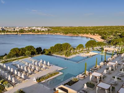 Hotelpool mit Liegestühlen und Blick auf einen bewaldeten Küstenabschnitt und den Strand.