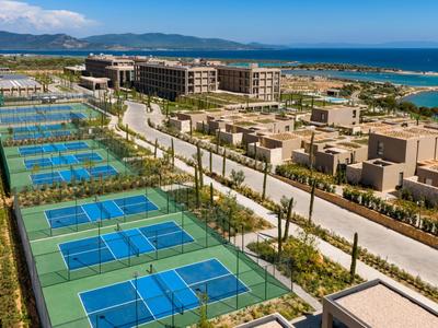 Mehrere blaue Tennisplätze neben modernen Hotelgebäuden mit Blick auf das Meer und Berge.