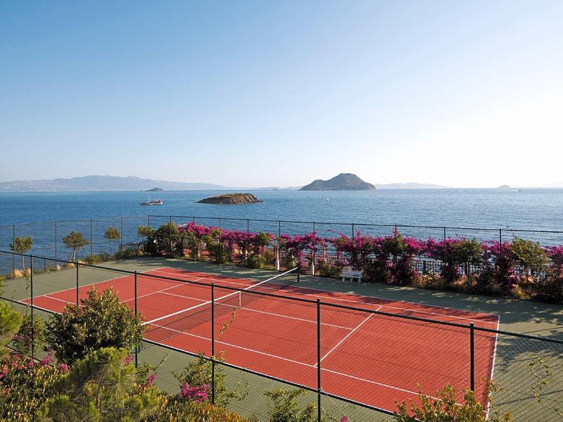 Ein roter Tennisplatz umgeben von grünem Buschwerk mit Blick auf das blaue Meer und kleine Inseln.