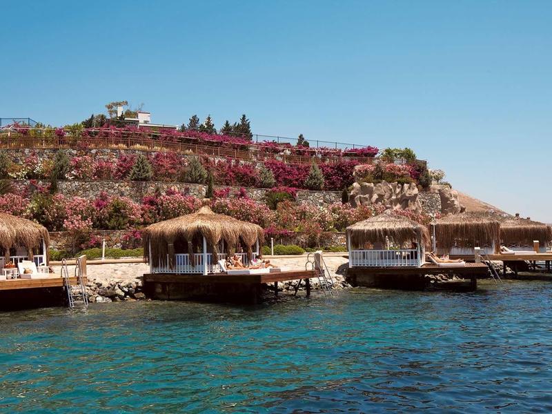 Klare blaue Küste mit Holzstegen und Strohhütten, im Hintergrund bunte Blumen und Häuser am Hang.