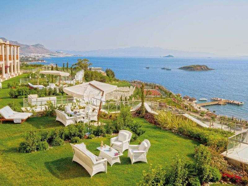 Grüne Gartenanlage mit weißen Gartenmöbeln, Blick auf blaue Meerwasserfläche und Inseln im Hintergrund.