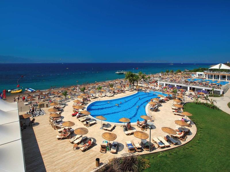 Large swimming pool with sun loungers and people by the sea under clear sky.