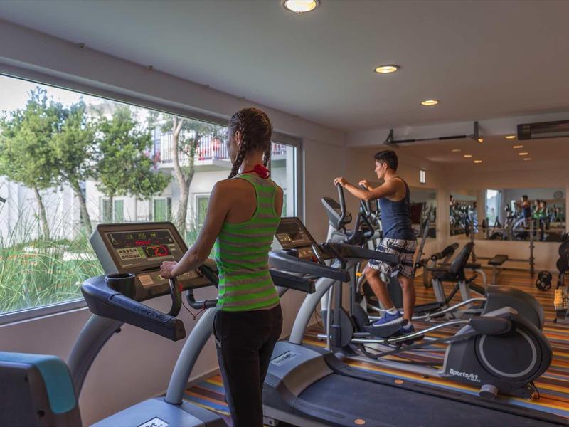 Two women exercise on cardio machines in a gym with large windows overlooking greenery.