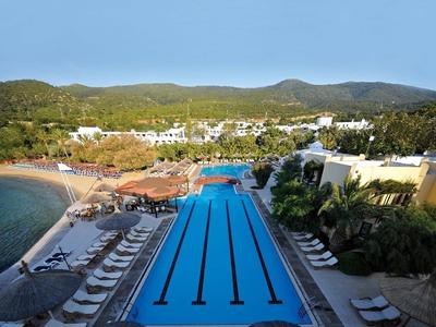 Piscine avec couloirs et chaises longues près de la mer, entourée d'hôtels et de collines verdoyantes.