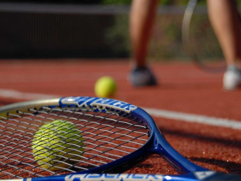 Close-up of a tennis racket and ball on a red tennis court.