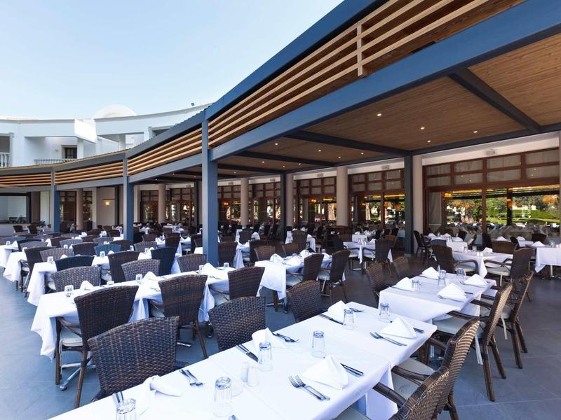 Large shaded outdoor restaurant with white tablecloths and many chairs under a modern wooden roof.