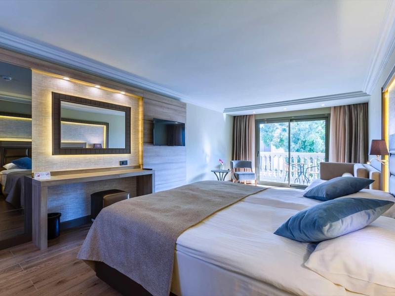 Modern hotel room with large bed, TV, desk, and balcony overlooking greenery.