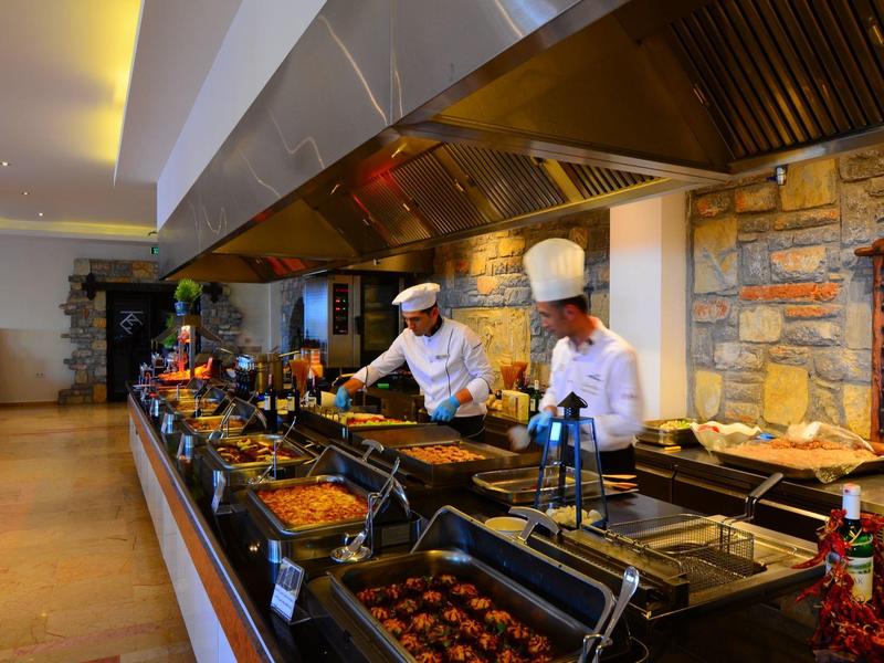 Große professionelle Küche mit Köchen, vielen Pfannen und warmem Licht an einer langen Arbeitsfläche.